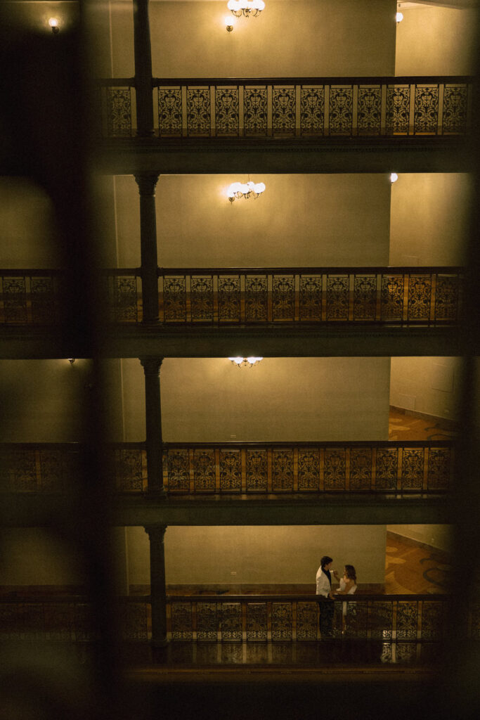 A couple standing on an indoor balcony of a hotel with three other floors above them. 