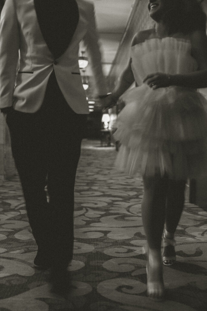 A wedding couple holding hands and walking down a hotel hallway. 