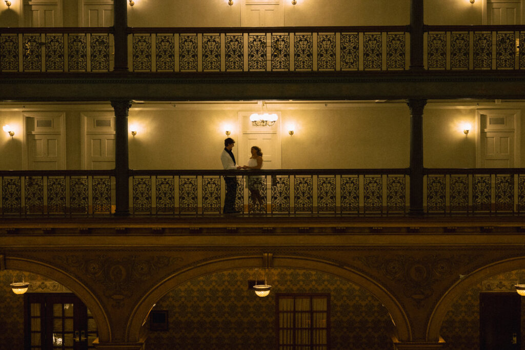 Image from across the lobby of a couple standing on an indoor balcony at a hotel 