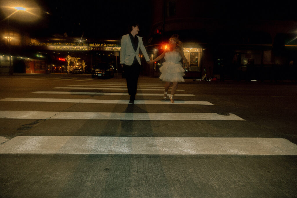 A newlywed couple holding hands and walking across a crosswalk 