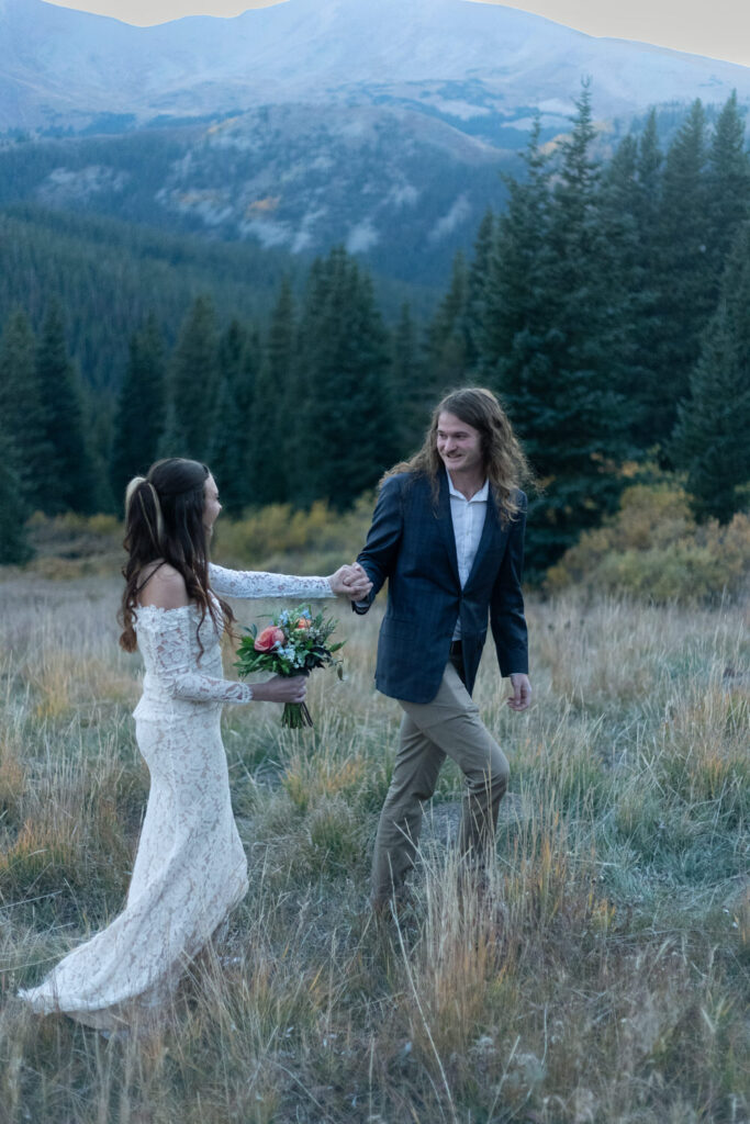 A couple holding hands and walking along a mountain while one of them holds a bouquet of flowers. 