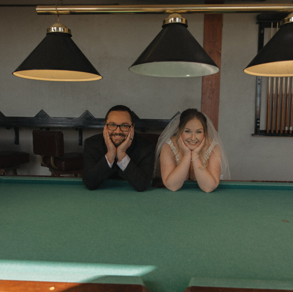 A wedding couple with their elbows on a pool table and their heads in their hands. 