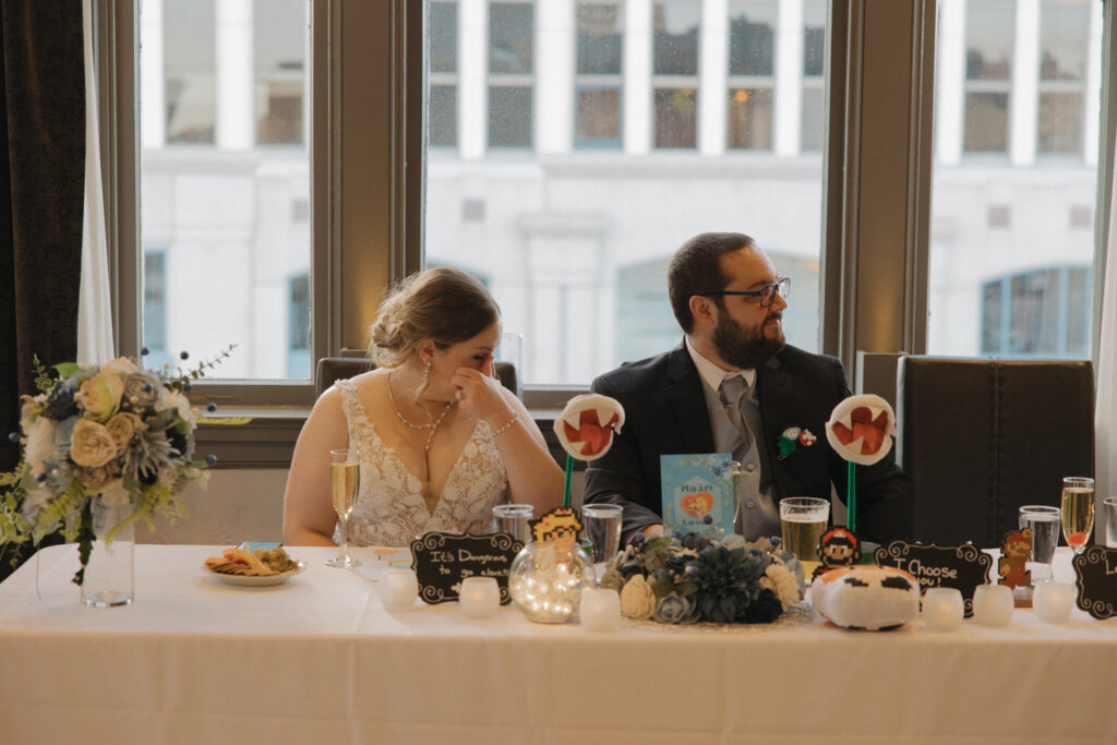 A wedding couple sitting at a table at their reception as one wipes away tears. 