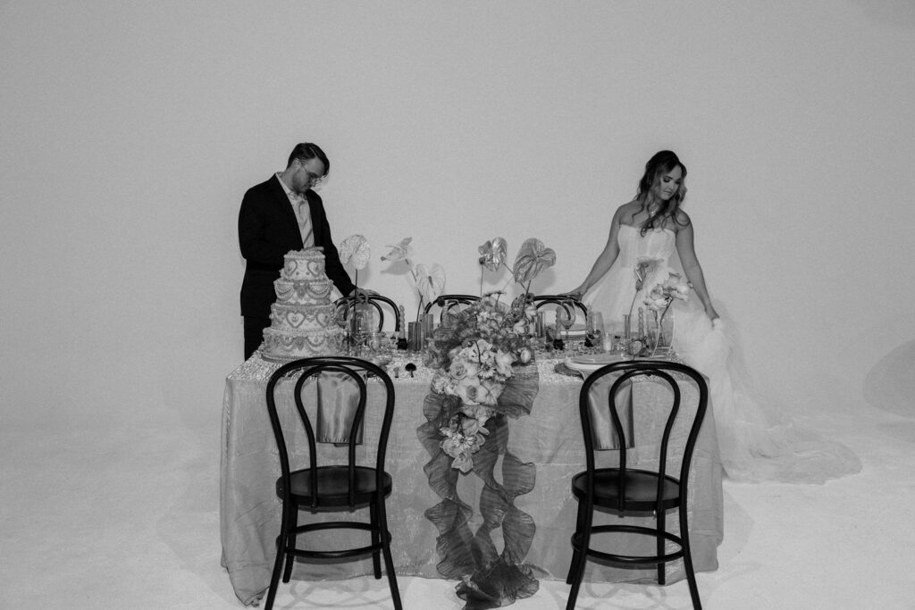 A couple standing on either side of a wedding reception table with a cake and decorations on it. 