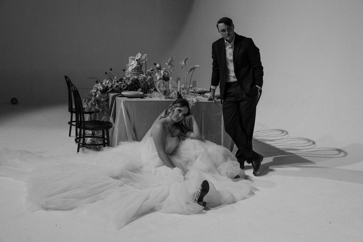 A black and white non-traditional wedding photos showing a person in a wedding dress sitting on the ground while their partner stands next to them