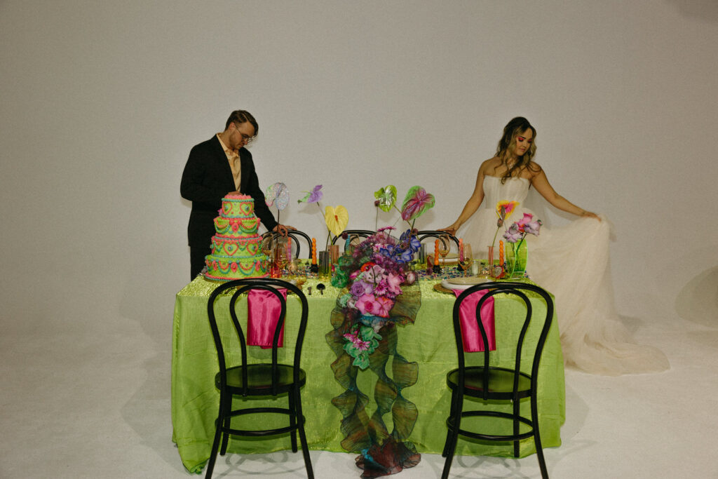 A person adjusting their wedding dress as their partner stands on the other side of a wedding reception table. 