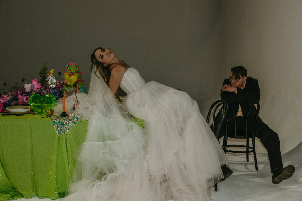 A person leaning back on a brightly decorated wedding reception table as their partner sits backwards in a chair next to them. 