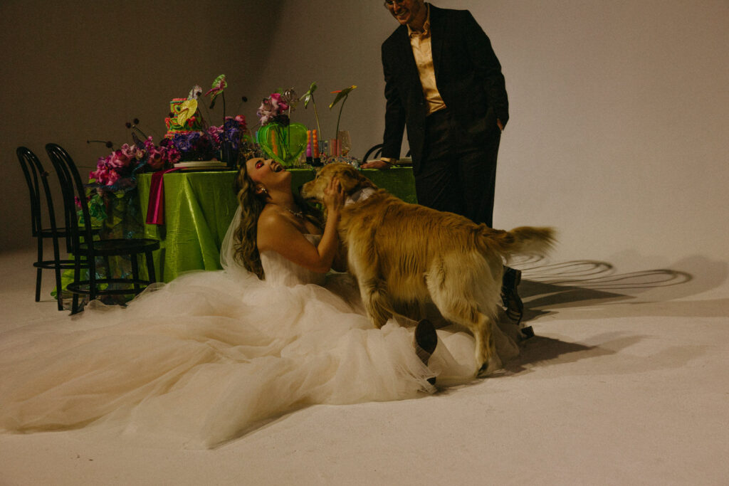 A person sitting on the floor in a wedding dress petting a dog as their partner stands next to them. 