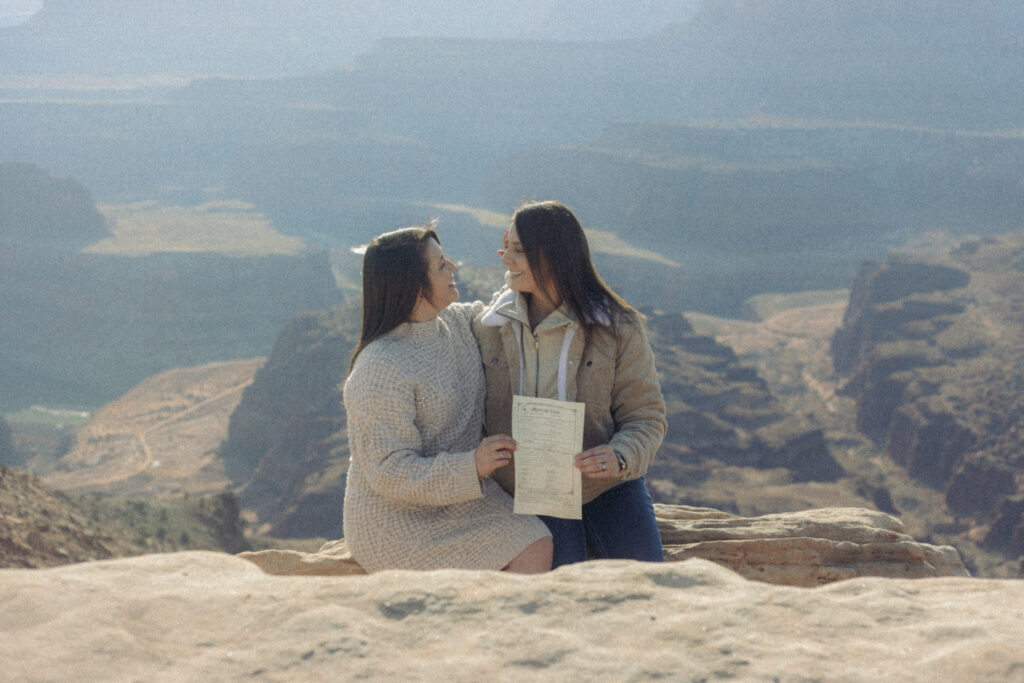 A couple sitting down on a rock holding out their marriage certificate 